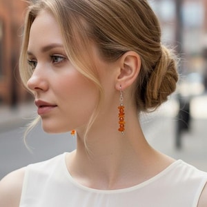May include: A pair of drop earrings featuring a vertical arrangement of small, vibrant orange beads suspended from a thin silver-toned metal wire. The earrings hang straight down, showcasing a series of rounded, textured orange elements.