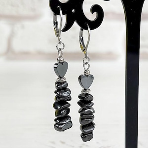 May include: A pair of dangle earrings with a heart-shaped top and a cascade of dark gray, irregularly shaped beads. The earrings are displayed on a black stand against a white brick wall and a white wooden surface. The earrings are made of hematite.