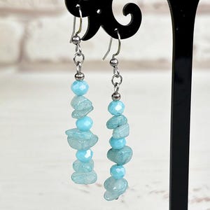 May include: A pair of dangle earrings with light blue beads and stones. The earrings are displayed on a black stand. The background includes a bouquet of yellow flowers and a white brick wall.