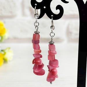 May include: A pair of dangle earrings featuring irregular, polished pink stone chips suspended from silver-toned metal hooks. Each earring consists of a vertical arrangement of multiple pink stone fragments, creating a textured, layered appearance that hangs from a small, clear crystal accent near the top of the hook.