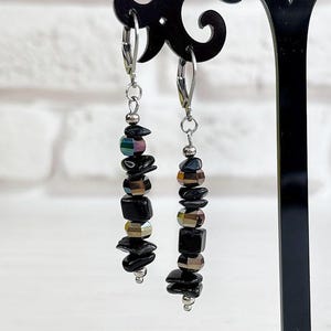 May include: Dangle earrings with black, iridescent, and metallic beads. The earrings are displayed on a black stand. The background is white and a brick wall.