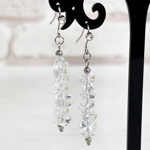 Puede incluir: Un par de pendientes colgantes con cuentas de cristal transparente facetadas ensartadas en alambres de metal plateado. Los pendientes cuelgan de ganchos plateados y se exhiben en un soporte para joyas negro, ornamentado y en forma de T.