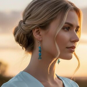 May include: A pair of dangle earrings featuring stacked, irregular turquoise-colored beads on a silver-toned wire. The earrings are worn by an individual with blonde hair styled in a low bun, wearing a light blue V-neck top against a sunset sky.