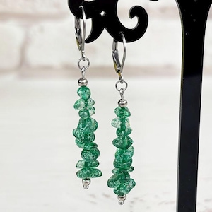 Może przedstawiać: Kolczyki wiszące z kaskadą małych, zielonych koralików o nieregularnym kształcie. Kolczyki zawieszone są na srebrnych haczykach i prezentowane na czarnym, dekoracyjnym stojaku. Tło obejmuje bukiet żółtych kwiatów i białą ceglaną ścianę.