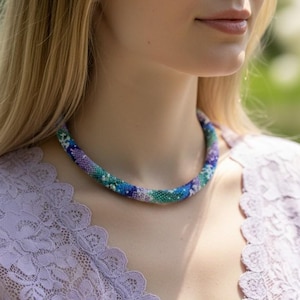 Può includere: Una collana di perline blu, verdi, viola e bianche. La collana è indossata su un top di pizzo lavanda. Le perline sono ben infilate, creando una forma liscia e arrotondata. Lo sfondo è un verde morbido e sfocato.