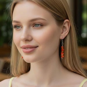 May include: Dangle earrings featuring small, irregular, bright orange coral-like beads strung on a thin silver-toned metal wire. The earrings hang from a simple silver-toned hook mechanism, creating a vertical, slender profile against the earlobe.
