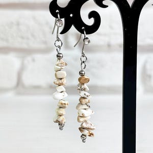May include: A pair of dangle earrings displayed on a black stand. Each earring features a silver chain with a cluster of small, irregular, off-white and tan stones. The earrings are set against a white brick wall and a bouquet of yellow flowers.