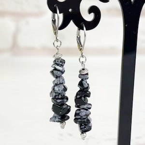 Może przedstawiać: Kolczyki wiszące z czarnymi i szarymi koralikami z kamienia. Kolczyki są prezentowane na czarnym stojaku. Kolczyki mają srebrne haczyki i są wykonane z naturalnych odłamków kamienia.