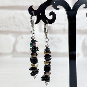 Può includere: Orecchini pendenti neri e argento con un mix di perline nere, iridescenti e color metallo. Gli orecchini sono esposti su un supporto decorativo nero su uno sfondo bianco e di mattoni.