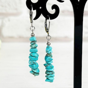 May include: Turquoise chip earrings hanging on a black display stand. The earrings are made of small, irregular turquoise stones strung together. The stand is on a white surface with a bouquet of yellow flowers in the background.