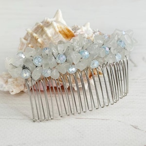 May include: A silver hair comb adorned with clear and light blue crystals and small flower-shaped accents. The comb is set against a white background with a seashell, suggesting a bridal or special occasion accessory.