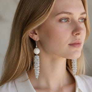 May include: A pair of long dangle earrings featuring a white spherical bead at the top, connected to a cluster of small, translucent, light-reflecting beads that form a tapered, textured fringe shape hanging vertically from the earlobe.