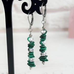 May include: Dangle earrings featuring green malachite and silver beads. The earrings are presented on a black stand. The design incorporates layered green stones and silver accents, creating a striking visual contrast.