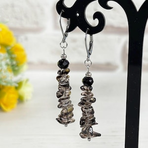 May include: A pair of dangle earrings displayed on a black stand. Each earring features a silver hook, a black bead, and a cascade of smoky quartz chips. The earrings are set against a backdrop of yellow flowers and a white brick wall.