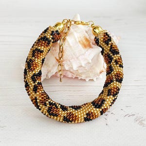 Può includere: Un braccialetto di perline con un motivo leopardato nei toni del marrone, nero e oro. Il braccialetto è appoggiato su una conchiglia. La chiusura e la catena sono color oro. Lo sfondo include un muro di mattoni bianchi e una scatola con la scritta "MADE IN UKRAINE".