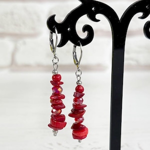 May include: A pair of dangle earrings with red beads and accents. The earrings are displayed on a black stand. The beads are a mix of shapes and sizes, creating a layered effect. The earrings have a silver-colored clasp.