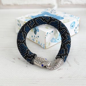 Può includere: Un bracciale flessibile di perline con un motivo geometrico blu scuro e nero. Il bracciale ha la forma di un serpente avvolto con una chiusura in metallo argentato che raffigura la testa e la coda, impreziosita da piccole pietre blu per gli occhi.
