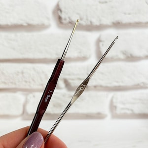 May include: Two crochet hooks, one with a brown handle and the other with a silver handle. The brown hook is labelled "1.0mm" and the silver hook is labelled "1.00mm".