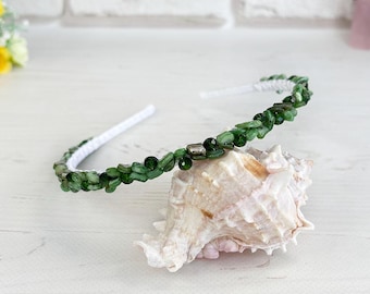 May include: A white headband adorned with small, faceted green gemstone beads and a single metallic silver spacer bead. The headband is resting on a large, textured, off-white seashell, showcasing a delicate and detailed construction.