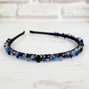 Puede incluir: Una diadema negra adornada con una variedad de cristales azules y transparentes. Los cristales varían en tamaño y tono, creando un efecto brillante. Un gran cristal negro está centrado en la diadema. El accesorio está sobre una superficie de madera blanca.