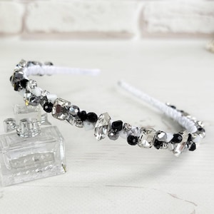 Puede incluir: Una diadema blanca adornada con cristales negros, transparentes y plateados. El accesorio para el cabello decorativo está adornado con piedras brillantes de varias formas y tamaños. Un frasco de perfume transparente está frente a la diadema.