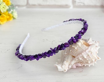 May include: A thin headband featuring a white base adorned with small, polished purple stone chips arranged in a continuous row along the top. The headband is curved and rests on a textured, light-coloured seashell.