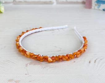 May include: A white headband adorned with numerous small, polished amber-coloured stone chips. The stones are arranged in a dense, continuous row along the outer curve of the flexible white band, creating a textured, warm-toned appearance.