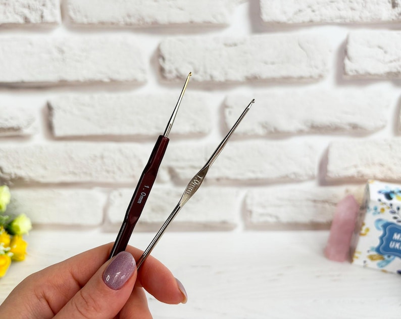 May include: Two crochet hooks, one with a brown handle and the other with a silver handle. The brown hook is labelled "1.0mm" and the silver hook is labelled "1.00mm".