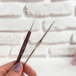 May include: Two crochet hooks, one with a red handle and the other with a silver handle. The red handled hook is labelled "1.0 mm" and the silver handled hook is labelled "1.00 mm".