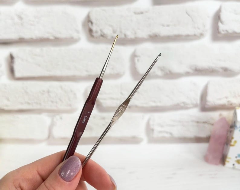 May include: Two crochet hooks, one with a red handle and the other with a silver handle. The red handled hook is labelled "1.0 mm" and the silver handled hook is labelled "1.00 mm".