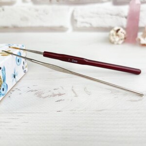 May include: Two crochet hooks, one silver and one red, on a white wooden surface. The red hook is labelled "1.0mm".