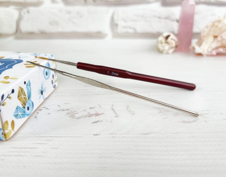 May include: Two crochet hooks, one silver and one red, on a white wooden surface. The red hook is labelled "1.0mm".