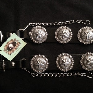 Puede incluir: Dos correas de cuero negro con hebillas de estrella plateadas y una cadena plateada. Las correas tienen una hebilla en un extremo y un anillo en D en el otro. Las correas están etiquetadas como "Cactus Mountain".