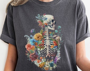 May include: A dark grey t-shirt featuring a floral skeleton design. The skeleton is surrounded by colourful flowers, including pink, blue, yellow and orange.