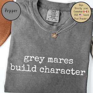 May include: A gray t-shirt with the text "grey mares build character" in white. The shirt has a crew neck and short sleeves. A silver necklace with a pendant is visible. The shirt is labeled "Comfort Colors".