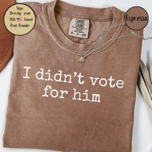 May include: A brown t-shirt with the text "I didn't vote for him" printed in white. The shirt is made of a soft, comfortable material and has a distressed look.