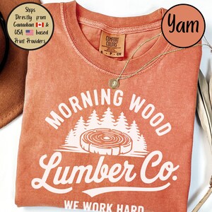 以下が含まれることがあります： 「Morning Wood Lumber Co. We Work Hard」と白で書かれた、焦げ茶色のTシャツ。グラフィックには、成長輪のあるスタイリッシュな木の切り株と、その背後に森の木々が描かれている。