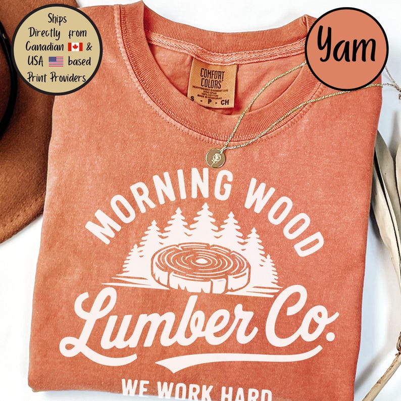 以下が含まれることがあります： 「Morning Wood Lumber Co. We Work Hard」と白で書かれた、焦げ茶色のTシャツ。グラフィックには、成長輪のあるスタイリッシュな木の切り株と、その背後に森の木々が描かれている。