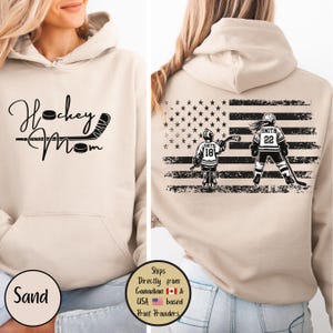 May include: Sand-colored hoodie with the words "Hockey Mom" in a script font. The back features a distressed American flag design with two hockey players, one with the number 18 and the other with the number 22.