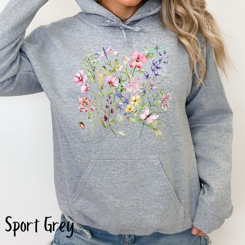 K&ouml;nnte beinhalten: Ein graumelierter Hoodie mit einem Blumenmuster mit rosa, gelben und violetten Blumen, gr&uuml;nen Bl&auml;ttern und Libellen. Der Hoodie hat eine Kapuze mit Kordelzug und eine K&auml;ngurutasche.