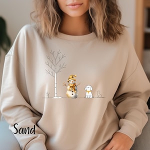 May include: A beige sweatshirt with a winter scene design. The design features a snowman, a dog, and a tree. The text "Sand" is printed on the bottom left corner of the sweatshirt.