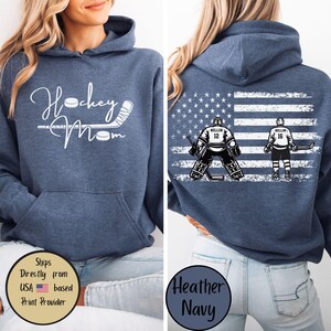 May include: Heather navy blue hoodie with the text "Hockey Mom" in white script on the front. The back features an American flag design with hockey players and the numbers 12 and 16. A cozy, casual top.