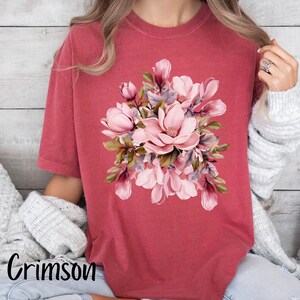 Peut inclure: Un t-shirt de couleur cramoisie avec un motif floral compos&eacute; de fleurs de magnolia roses et de feuilles vertes.