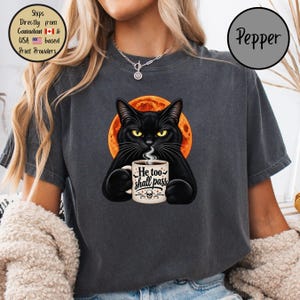 May include: A charcoal gray t-shirt featuring a graphic of a black cat with yellow eyes holding a white coffee mug. The mug displays the text He too shall pass with a small skull icon. Behind the cat is a large, bright orange full moon.