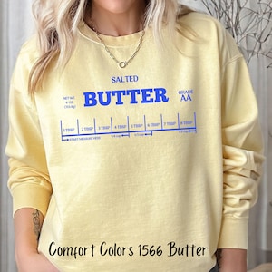 May include: A yellow sweatshirt with the text "SALTED BUTTER GRADE AA" and a measurement guide for tablespoons of butter. The text "Comfort Colors 1566 Butter" is printed on the bottom of the sweatshirt.