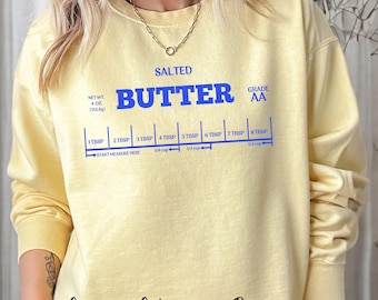 May include: A yellow sweatshirt with the text "SALTED BUTTER GRADE AA" and a measurement guide for tablespoons of butter. The text "Comfort Colors 1566 Butter" is printed on the bottom of the sweatshirt.