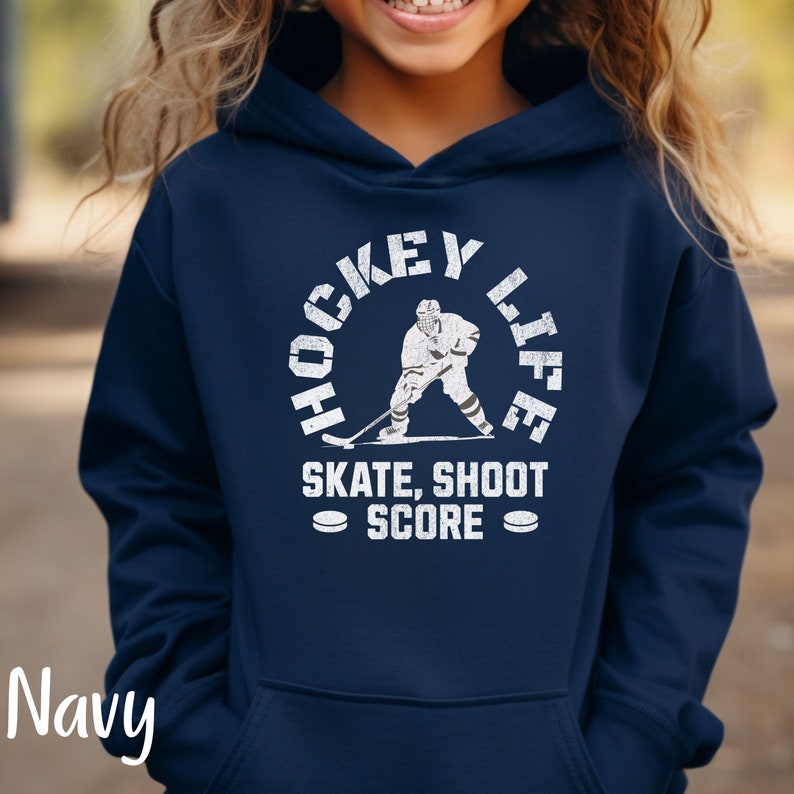 Op de afbeelding: Donkerblauwe hoodie met een witte afbeelding van een hockeyer in een cirkel met de tekst "Hockey Life" en "Skate, Shoot Score" eronder.
