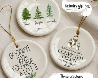 May include: Three round white ceramic ornaments with gold hanging strings. One features three green trees with the names Fraser, Douglas, and Tamanack and the year 2026. Another shows three snowmen with text reading Another Christmas with a CONVICTED FELON as President 2026. The third says GOODBYE 2026 YOU WERE....... A LOT !! with a small holly sprig.