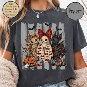 Peut inclure: Un t-shirt gris anthracite avec un motif de trois chauves-souris de dessin animé aux ailes déployées. Les chauves-souris sont ornées d'imprimés léopard, de cerises rouges et de crânes. Le design inclut une citrouille, des crânes et des fioles.