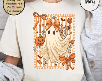 May include: Ivory-colored t-shirt featuring a Halloween ghost design. The ghost is adorned with a bow and surrounded by pumpkins, bats, and spiderwebs. The design is framed by orange stripes and decorative elements. The shirt also has the word "Ivory" printed on it.